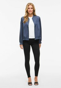 Giacca bomber in satin blu navy con zip frontale, polsini e orlo a coste, abbinata a una maglietta bianca e leggings neri, con scarpe nere con la punta aperta.