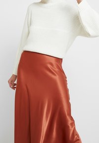 Pull en tricot blanc avec un col montant, associé à une jupe en satin couleur rouille qui tombe avec fluidité, créant une silhouette élégante.