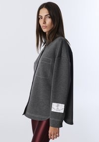 Felpa grigia oversize con colletto a punta, tasca a toppa e dettagli con etichetta bianca sulla manica; texture liscia con cuciture a contrasto.