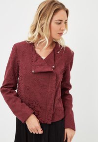 Chaqueta corta burdeos con una intrincada textura floral, cierre de botón a presión y un diseño de cuello diagonal. Mangas largas y sutiles detalles de fruncido.