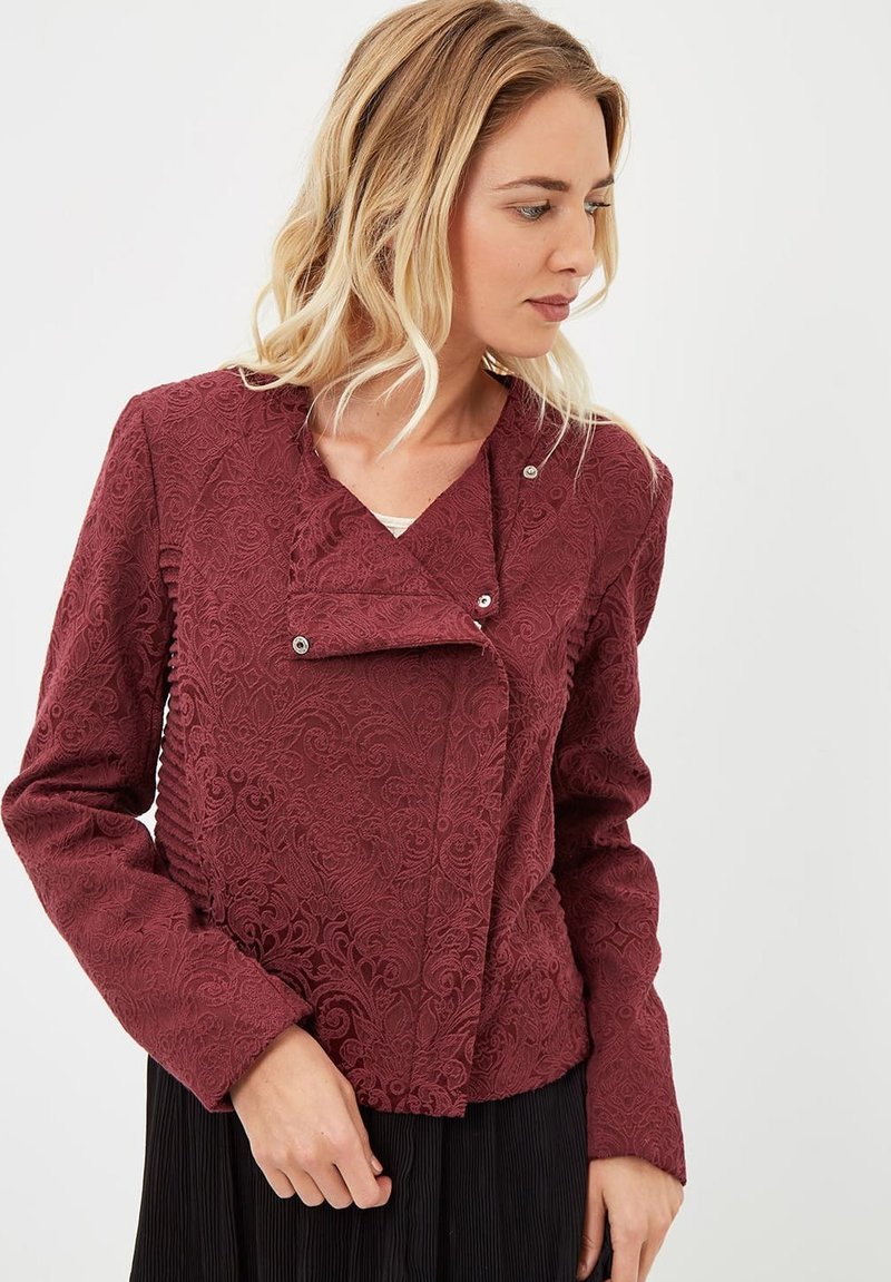 Chaqueta corta burdeos con una intrincada textura floral, cierre de botón a presión y un diseño de cuello diagonal. Mangas largas y sutiles detalles de fruncido.