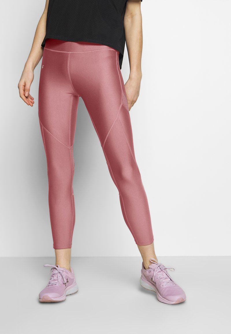 Personne portant des leggings roses brillants, des chaussures de sport roses et un t-shirt noir, debout devant un fond clair uni.