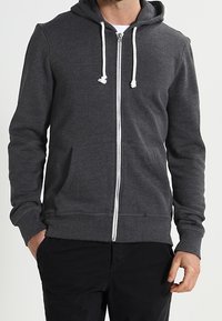 Sudadera con capucha gris con cremallera, confeccionada en tela suave y texturizada. Cuenta con cremallera frontal, cordones blancos y bolsillos laterales. Diseño ligero y casual.