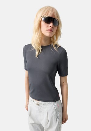 Femme blonde portant des lunettes de soleil noires surdimensionnées, un haut ajusté à manches courtes gris foncé et un pantalon blanc ample, sur fond uni.