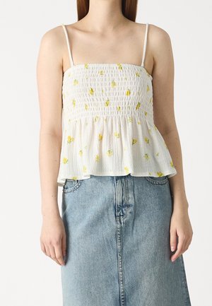 Kvinde iført en hvid smocksømme ærmeløs top med gult blomsterprint og en lyseblå denim nederdel mod en ensfarvet baggrund.