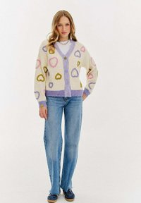 Pletený kardigan v krémové barvě s barevnými vzory srdcí; fialový lem a zapínání na knoflík. Kombinovaný se světlými modrými džíny a námořnickými botami.