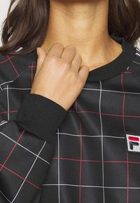 Černá mikina se vzorem mřížky v bílé a červené. Hladké manžety a límec. Obsahuje malou nášivku s logem v modré, bílé a červené.