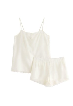 Kvinde står barfodet iført hvid camisole og matchende shorts med blondedetaljer, smilende med håret samlet i en hestehale.