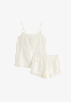 Kvinde står barfodet iført hvid camisole og matchende shorts med blondedetaljer, smilende med håret samlet i en hestehale.