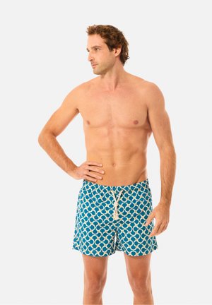 Paidaton mies, jolla on kiharat hiukset ja siniset kuvioidut uimashortsit valkoisella nyörillä, seisoo käsi lantiolla ja katsoo vasemmalle.