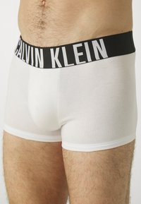 Calvin Klein hvite boxertruser med svart midjebånd som har hvit tekst. Glatt bomullsmateriale, tettsittende passform og midt på låret lengde.