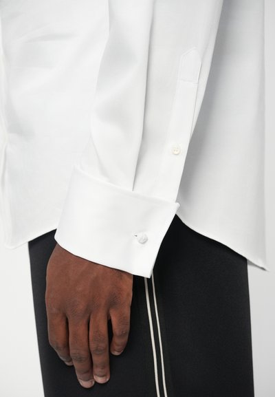 White long-sleeve shirt with a buttoned cuff, featuring a smooth texture and a clean design. Hand resting on dark pants with side stripes.