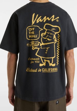 Zwart Vans T-shirt met gele afbeelding van een chef-kok die een schoen vasthoudt, tekst luidt "OFF THE WALL," "Custommade for YOU," en "Baked in CALIFORNIA."