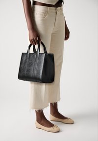 Zwarte leren tote bag met twee handvatten, met een textuur aan de oppervlakte en een reliëf logo. Gekoppeld aan crèmekleurige cropped broeken en beige flats.