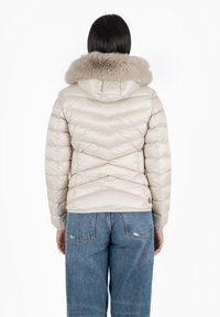 Giacca puffer beige chiaro con cappuccio, dotata di un colletto in pelliccia sintetica, cuciture a zig-zag diagonali e texture trapuntata. Abbinata a jeans blu.