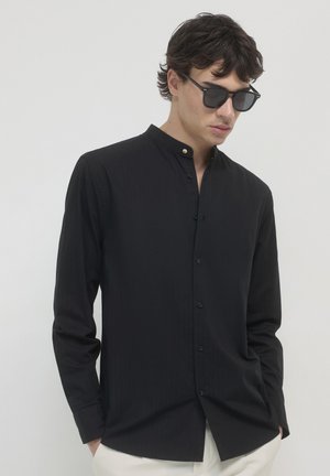 Schwarzes Langarm-Hemd mit Knopfleiste und Stehkragen, versehen mit dezenten vertikalen Streifen, kombiniert mit Sonnenbrille und hellen Stoffhosen.