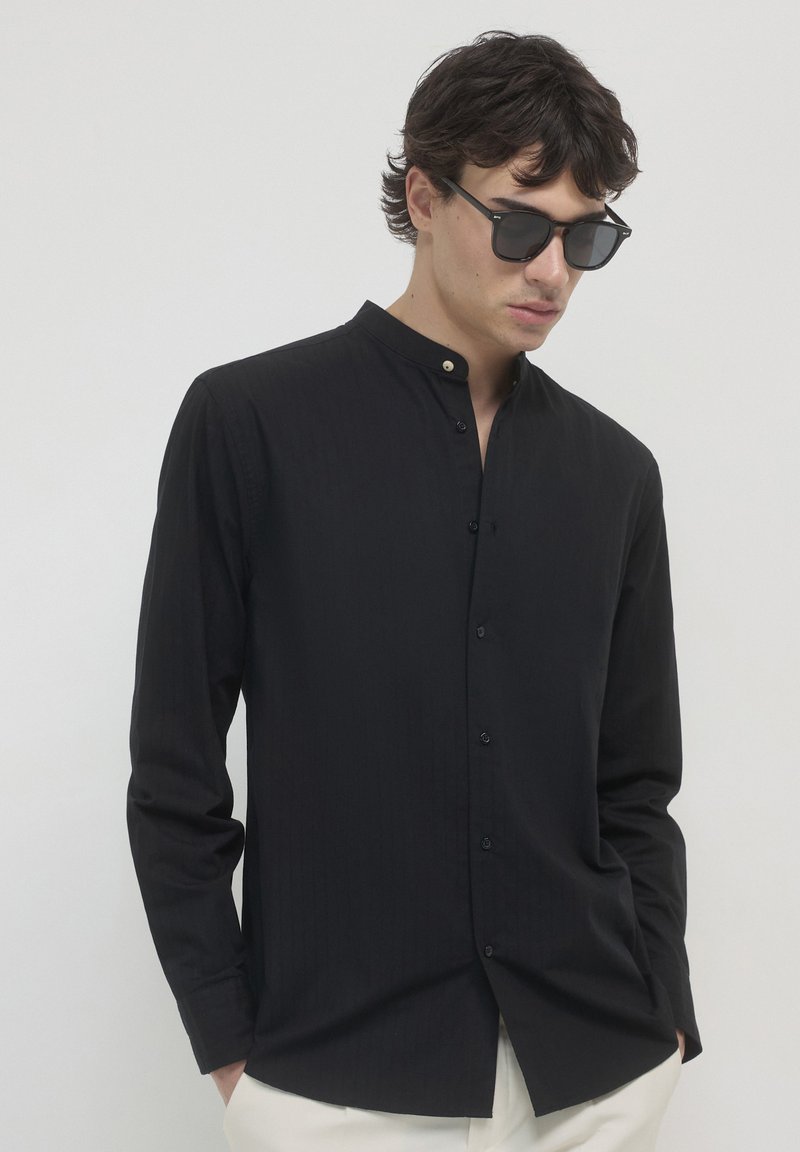 Schwarzes Langarm-Hemd mit Knopfleiste und Stehkragen, versehen mit dezenten vertikalen Streifen, kombiniert mit Sonnenbrille und hellen Stoffhosen.