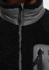 Chaqueta de forro polar negra con detalles de nailon gris, cuello alto, cremallera frontal y bolsillo con cremallera. Superficie texturizada y tela duradera.