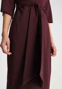 Robe bordeaux avec une texture douce, taille cintrée nouée d'un nœud en tissu, et manches courtes. Présente une coupe droite jusqu'aux genoux.