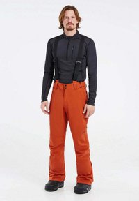 Orange vattentäta skidbyxor med justerbara hängslen, avsmalnande ben och svarta högstövliga snöskor. Lager på med en svart långärmad tröja.