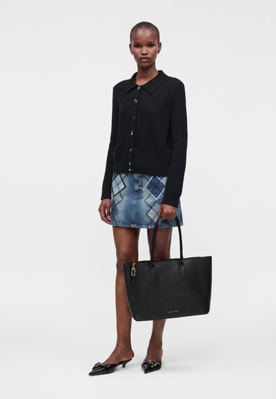 Femme portant un cardigan noir boutonné et une jupe en jean bleue à motifs, tenant un grand sac à main noir et portant des chaussures noires pointues.