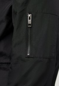 Svart tygjacka med en sidficka som har en synlig metallblixtlås, slät textur och en snygg, minimalistisk design.