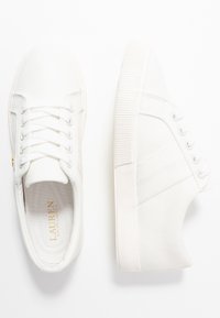 Zapatillas de lona blancas con una suela de goma texturizada, seis ojetes y un sutil logo en la plantilla. Presentan líneas simples y limpias.