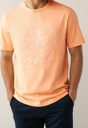 Mand iført en ferskenfarvet t-shirt med et billede af en palme og sol og teksten "Enjoy the waves" og "Surf Up" med hvide bogstaver.