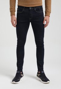 Hombre vestido con jeans ajustados azul oscuro, camisa de manga larga color beige y zapatillas en blanco y negro, de pie sobre un fondo blanco.