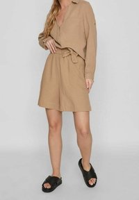 Béžový dvoudílný outfit sestávající z košile s dlouhým rukávem a přední kapsou a kraťasů s uvázaným pasem. V kombinaci s černými sandály na klínu.