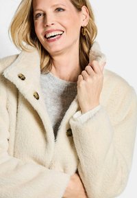 Crème-kleurige, gestructureerde jas met een hoge kraag en drie grote knopen. Gecombineerd met een lichtgrijs gebreid sweater. Zacht, pluizig materiaal.