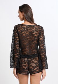 Blusa de encaje negro con mangas largas acampanadas y detalles transparentes, combinada con pantalones cortos de encaje negro a juego, con un patrón floral.