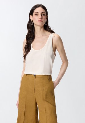 Mujer con cabello largo y oscuro que lleva una camiseta blanca sin mangas y pantalones de talle alto amarillos mostaza, de pie con una mano en el bolsillo.