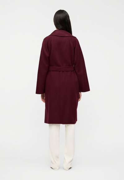 Μπουφάν από βελούδινο μάλλινο ύφασμα σε μπορντό χρώμα, με φαρδιά μανίκια, ζώνη στη μέση και γιακά. Φορεμένο πάνω από ανοιχτόχρωμο παντελόνι, με ομαλή υφή.