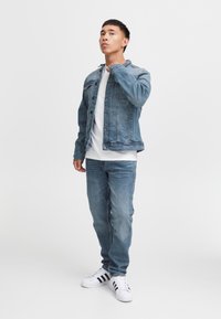 Ljusblå denimjacka över en vit t-shirt, kombinerad med ljusblå jeans och svartvita sneakers. Modell står i en neutral miljö.