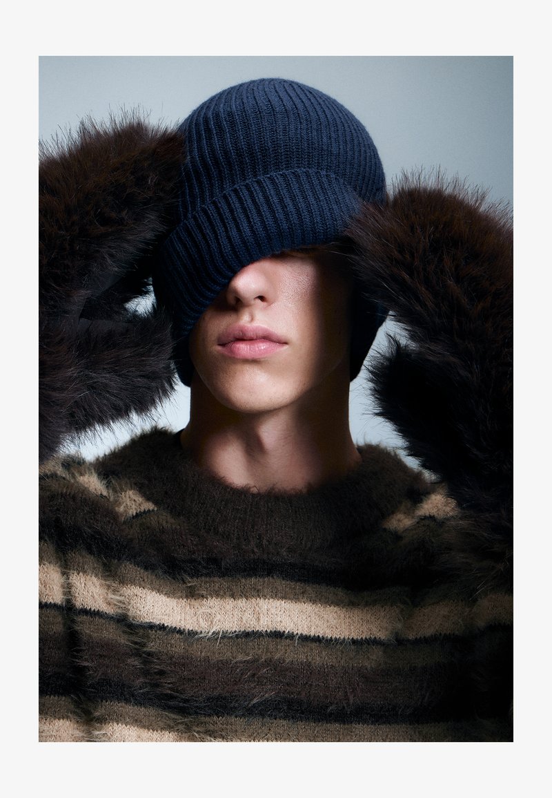 Osoba nosząca granatową dzianinową czapkę, naciągniętą na oczy, oraz puszysty, w prążki brązowy sweter, poprawiająca czapkę w futrzastych rękawiczkach.