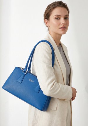 Donna in completo beige che tiene una borsa strutturata in pelle blu con doppi manici sulla spalla, su uno sfondo uniforme.