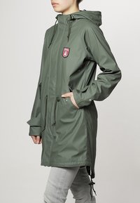 Personne portant un imperméable à capuche vert olive avec un écusson sur la poitrine, les mains dans les poches, portant un pantalon gris clair, debout de profil.