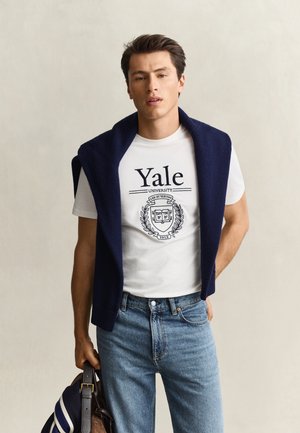 Jeune homme portant un t-shirt blanc de l'université de Yale et un jean bleu, avec un pull bleu marine posé sur les épaules, tenant un sac dans une main.