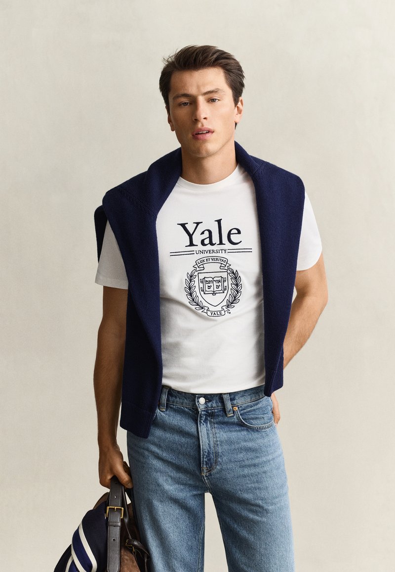 Jeune homme portant un t-shirt blanc de l'université de Yale et un jean bleu, avec un pull bleu marine posé sur les épaules, tenant un sac dans une main.
