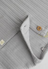 Chemise grise à côtes avec détails de boutons, présentant une texture de tissu douce et une petite étiquette de logo sur le côté gauche.
