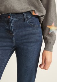 Jean en denim bleu foncé avec une texture lisse, doté d'un bouton à l'avant et de poches. Associé à un pull gris avec des accents étoilés colorés.