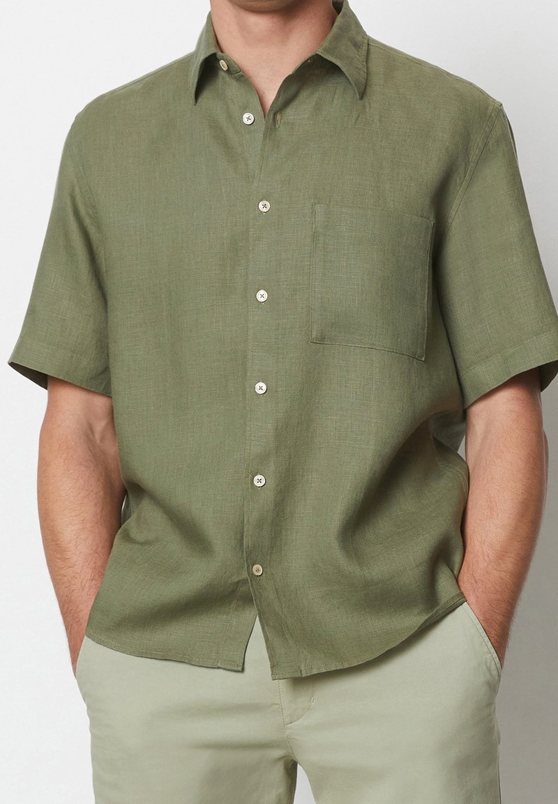 Homme portant une chemise à manches courtes vert olive avec une poche poitrine, associée à un pantalon beige clair, mains dans les poches.
