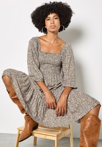 Florales Midikleid mit quadratischem Ausschnitt, Puffärmeln und geraffter Textur in Grün mit rosa und weißen Blumen; kombiniert mit braunen kniehohen Stiefeln.