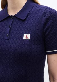 Μπλε σκούρο πόλο μπλουζάκι με πλέξη καλωδίου, γιακά, τρία μαύρα κουμπιά, λευκή ρίγα στην μανσέτα του μανικιού και λογότυπο Calvin Klein στο στήθος.