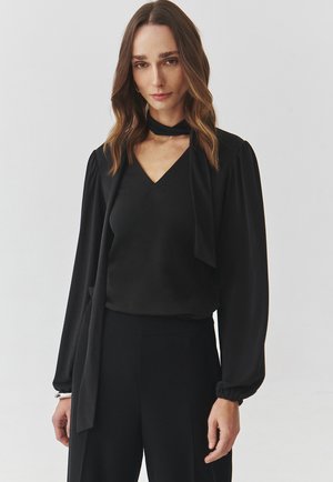 Black blouse with a V-neck and long, puffed sleeves. Features a tie around the neck and smooth fabric. Paired with black wide-leg pants.