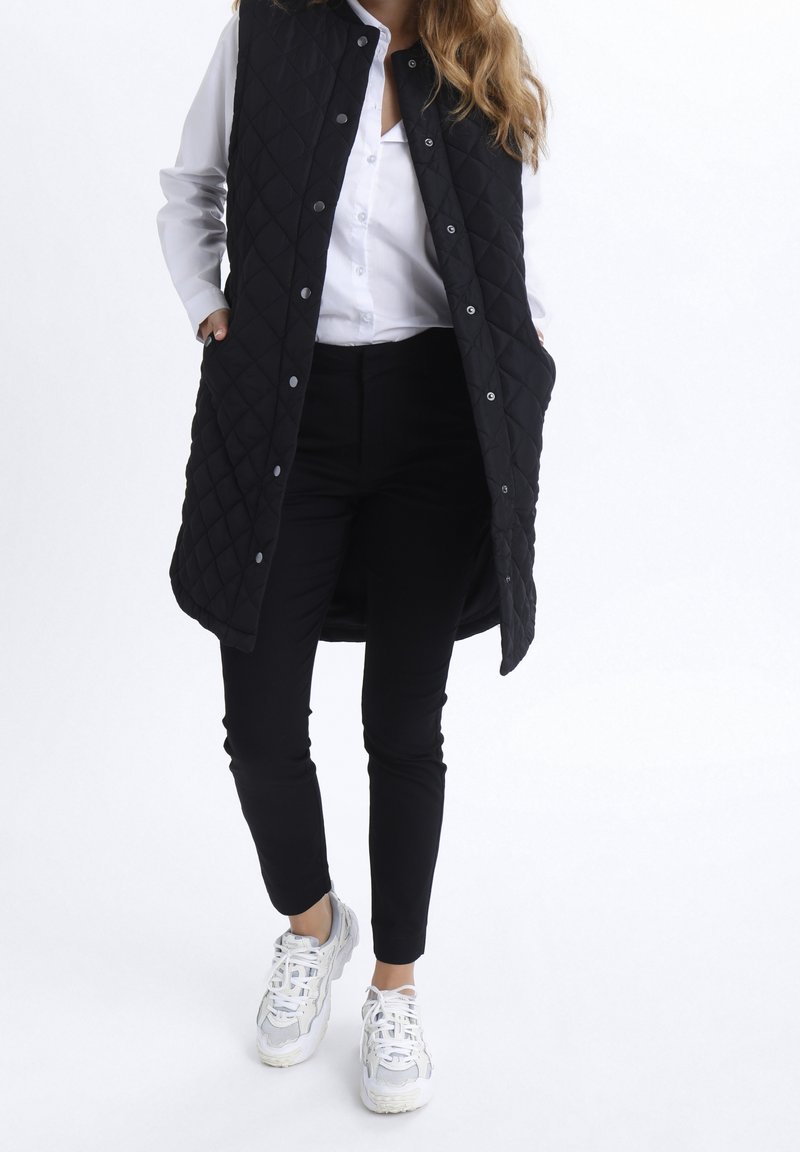 Femme portant une longue veste matelassée noire, une chemise blanche boutonnée, un pantalon noir et des baskets blanches, debout les mains dans les poches de la veste.