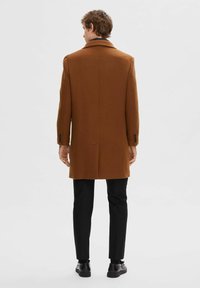 Brun ullfrakk med spiss krage, enkelt ventil, og tre knapper på ermene, kombinert med svarte bukser og sko.