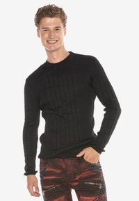 Schwarzer, gerippter Langarmpullover mit Rundhalsausschnitt und strukturiertem Stoff. Getragen mit roten Hosen, die metallische Akzente und Reißverschlüsse aufweisen.