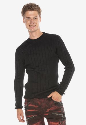 Schwarzer, gerippter Langarmpullover mit Rundhalsausschnitt und strukturiertem Stoff. Getragen mit roten Hosen, die metallische Akzente und Reißverschlüsse aufweisen.